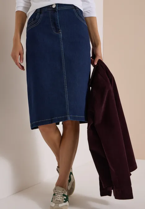 Une femme porte une jupe en jean mi-longue et tient une veste bordeaux.