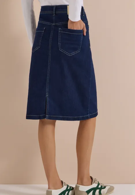 Jupe en jean bleu foncé mi-longue fendue au dos, portée avec des baskets blanches à bandes vertes.