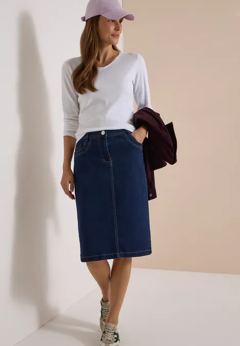 Femme en tee-shirt blanc à manches longues, jupe en jean mi-longue, baskets beiges et casquette rose, tenant une veste bordeaux.