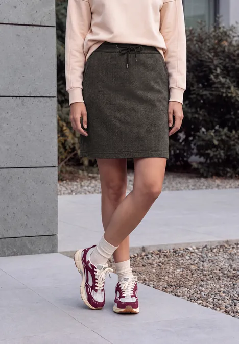 Vrouw staat buiten bij een gebouw, met grijze rok, roze trui en witte sneakers met paarse details.
