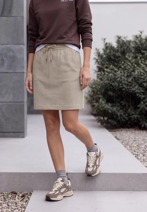 Vrouw in bruin sweatshirt, beige rok en sneakers loopt op een trap voor een modern gebouw met planten.