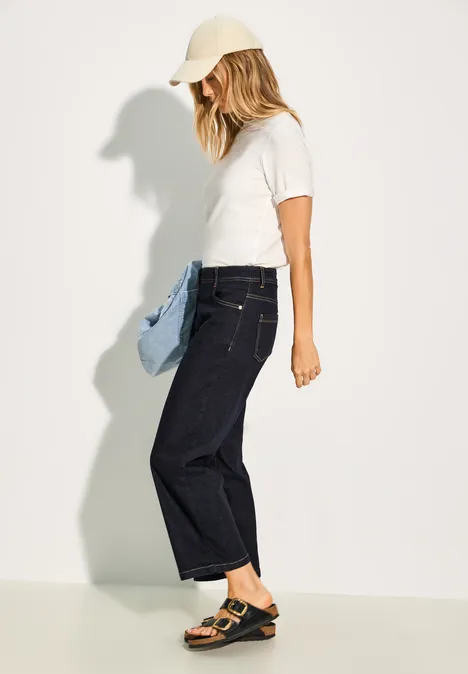 Femme en t-shirt blanc, jean foncé, casquette et sandales, tenant une veste bleue.