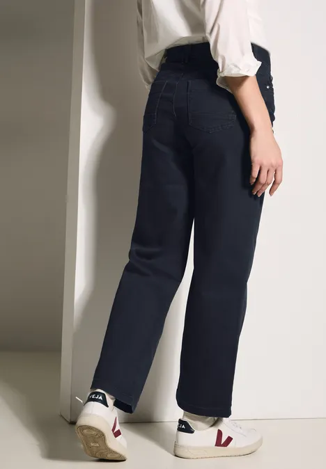 Femme en jean bleu foncé, chemise blanche et baskets blanches près d’un mur.