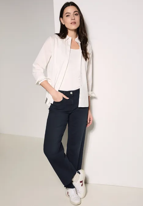 Femme en chemise blanche, top blanc et pantalon foncé, appuyée contre un mur.