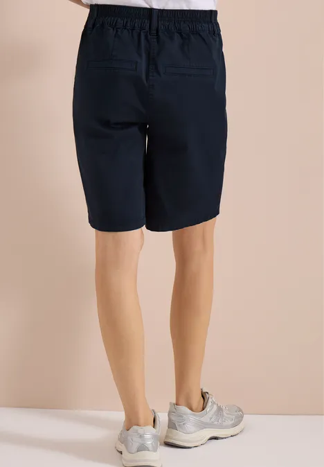 Achteraanzicht van persoon in donkerblauwe knielange short en lichte sneakers voor beige achtergrond.