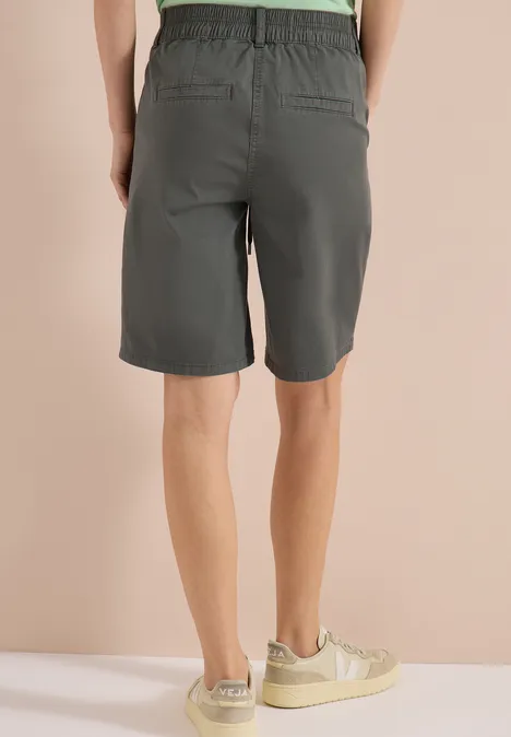 Achteraanzicht van persoon in grijze korte broek en lichte sneakers tegen een beige achtergrond.