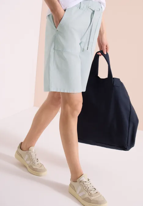 Een persoon draagt lichtgroene shorts, beige sneakers en houdt een grote zwarte tas vast.