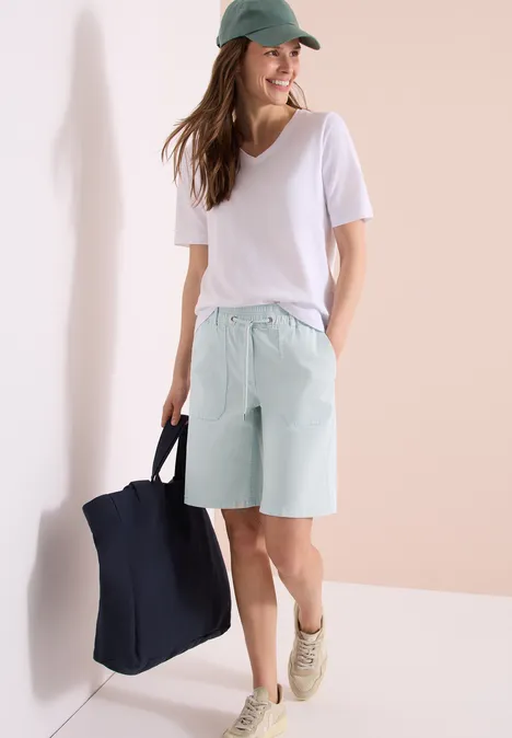 Vrouw in wit T-shirt, mintgroene bermudashorts en pet met een grote tas.