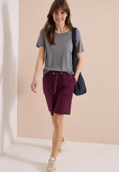 Een vrouw in grijs t-shirt, wijnrode bermuda's en sneakers, loopt met een blauwe schoudertas.