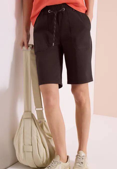 Een persoon draagt zwarte bermudashorts, lichte sneakers en houdt een beige tas vast.