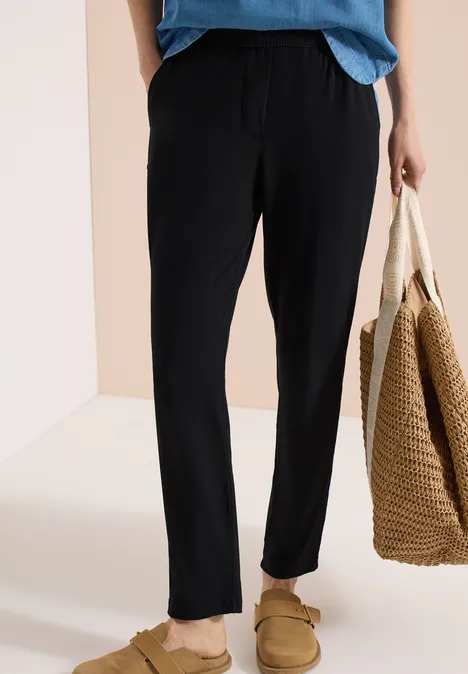 Person trägt schwarze, lässige Hose und Hausschuhe, hält eine geflochtene Tasche.