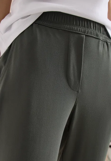 Nahaufnahme einer Person in dunkelgrüner Hose mit elastischem Bund und Seitentasche, kombiniert mit weißem Oberteil.