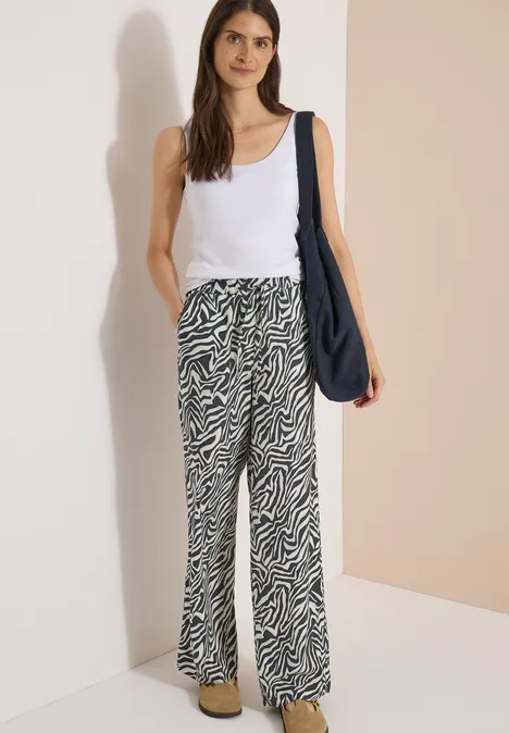 Pantalon Straight Leg avec imprimé zèbre palm breeze green Femme en débardeur blanc, pantalon large à motif zébré et sac bandoulière bleu marine, debout devant un mur beige.