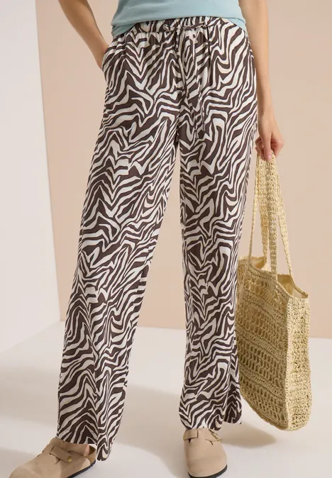 Pantalon Straight Leg avec imprimé zèbre brown cacao Femme porte un pantalon large imprimé zèbre noir et blanc, un top clair, un sac en osier et des chaussures plates.