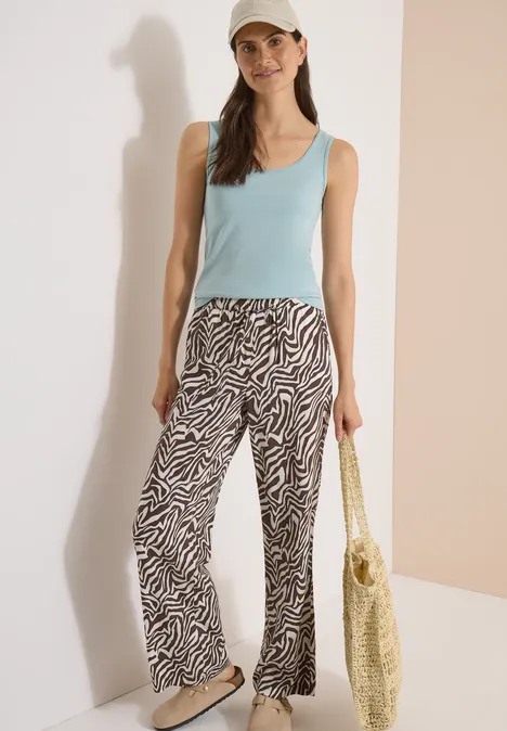 Pantalon Straight Leg avec imprimé zèbre brown cacao Femme en débardeur bleu clair, pantalon zèbre et casquette, portant un sac tressé devant un fond neutre.