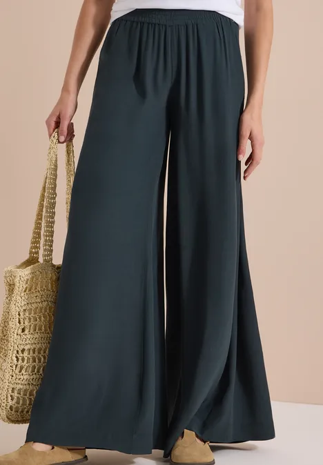 Vrouw draagt wijde, donkergroene broek met hoge taille, gecombineerd met strooien tas en beige schoenen.