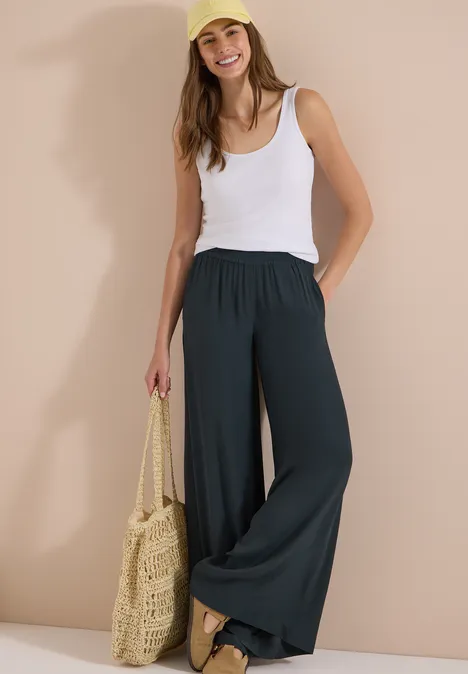 Vrouw in witte tanktop, wijde zwarte broek en rieten tas voor een casual zomerse stijl.