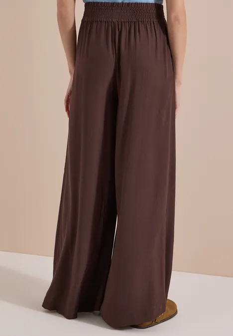 Rugaanzicht van persoon in een brede, bruine broek met elastische taille, vloeiende stof.