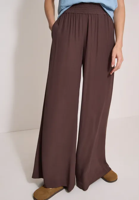 Wijdvallende bruine hoge taille broek met zakken, gecombineerd met een blauw topje en bruine schoenen voor een lichte achtergrond.