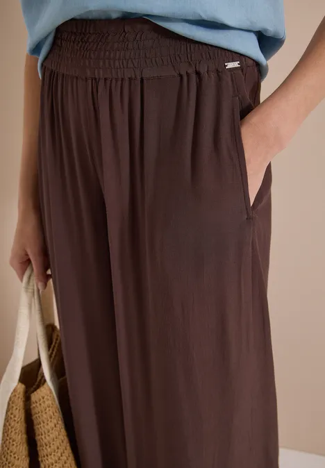 Close-up van een losse, bruine broek met elastische taille en zijzakken, hand in zak en rieten tas.