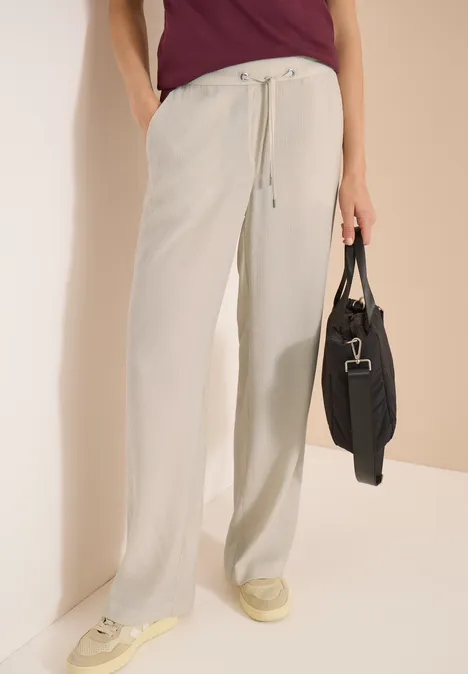 Personne porte un pantalon large en lin clair à cordon, des baskets crème et tient un sac noir.