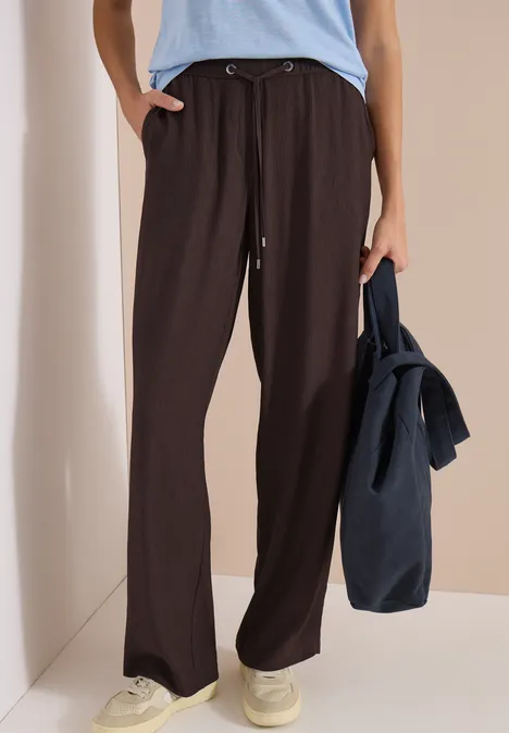 Frau trägt weite, dunkelbraune Hose, hellblaues Oberteil, weiße Sneaker, hält lässig eine dunkelblaue Tasche.