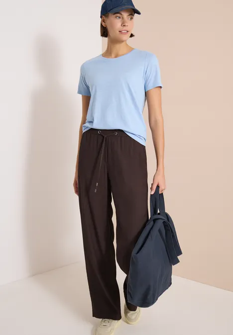 Eine Person in hellblauem T-Shirt und brauner Hose, hält eine dunkelblaue Tasche.