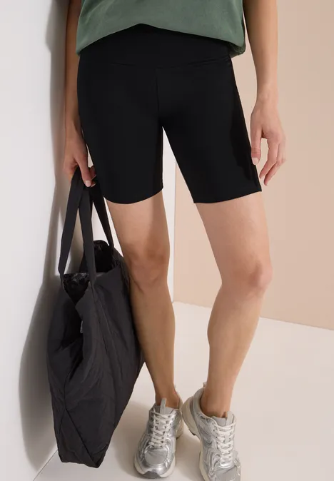 Vrouw met zwarte fietsshorts, groen shirt, zilveren sneakers en een grote gewatteerde zwarte tas.