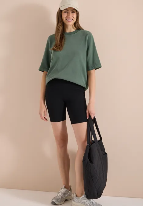 Vrouw met groen t-shirt en zwarte fietsshorts, met een grote zwarte tas voor een beige muur.
