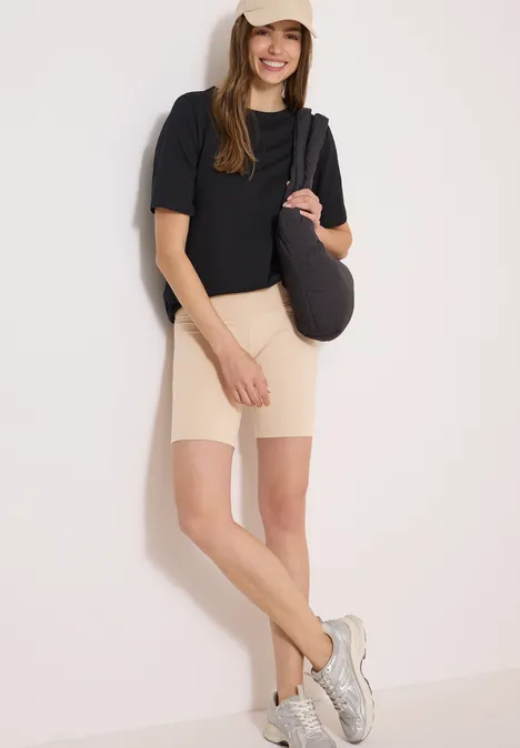 Frau in schwarzem T-Shirt, beigen Radlerhosen und weißen Sneakern, mit grauer Schultertasche vor heller Wand.