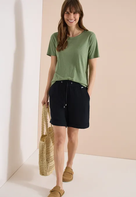 Frau in grünem T-Shirt und schwarzen Shorts, mit Strohtasche und Slippern in minimalistischem Setting.