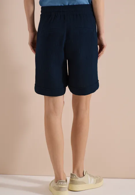 Ein Mann steht von hinten in dunkelblauen, knielangen Shorts und beigen Freizeitschuhen.