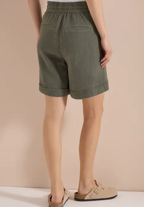 Rückansicht einer Frau in khaki Shorts und beigen Pantoletten vor schlichtem Hintergrund.