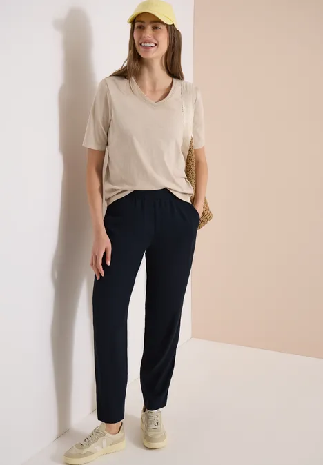 Vrouw leunt tegen een muur, draagt een beige t-shirt, donkerblauwe broek en beige sneakers.