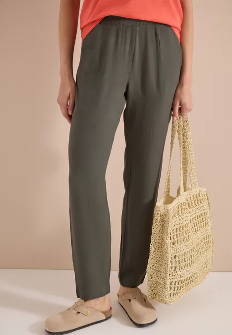 Iemand draagt olijfgroene joggingbroek, beige instapschoenen en houdt een gevlochten strozak vast.