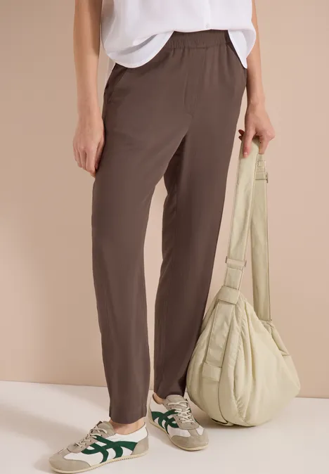 Vrouw draagt losse bruine broek, wit shirt, groene sneakers en een lichtgroene, gewatteerde schoudertas.