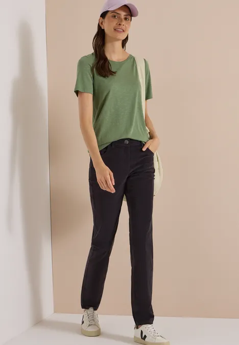 Femme en t-shirt vert, jean foncé, casquette beige et baskets blanches devant un mur beige.