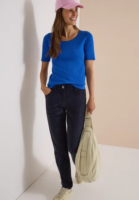 Eine Frau trägt ein blaues T-Shirt und eine rosa Kappe, hält eine helle Jacke vor neutralem Hintergrund.