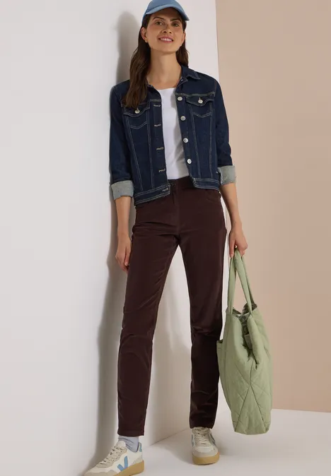 Mid Waist Slim Leg Hose im Slim Fit truffle brown Eine Frau in Jeansjacke, weißem Top, brauner Hose und Cap lehnt mit grünem Beutel an einer Wand.