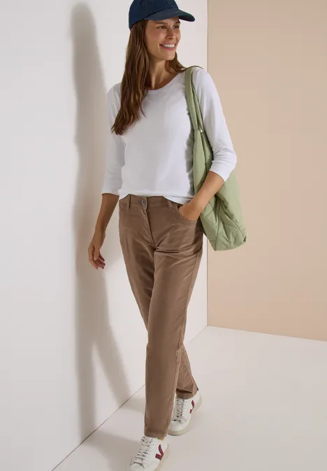 Frau mit Baseballcap, weißem Langarmshirt, braunen Cordhose, weißen Sneakern und grünem Shopper vor heller Wand.