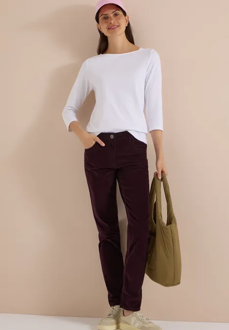 Femme en tee-shirt blanc à manches longues, casquette rose et pantalon bordeaux, avec un sac vert devant un mur beige.