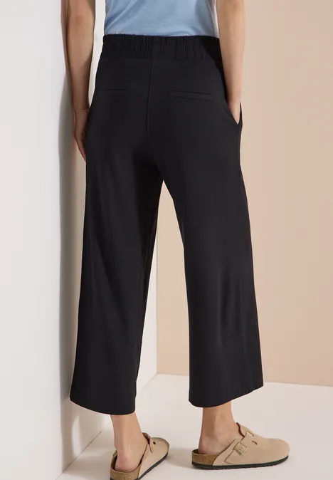 3/4 Wide Leg Hose im Casual Fit Black Frau von hinten in schwarzer, weiter, knöchellanger Hose mit Taschen, kombiniert zu beigen Clogs.