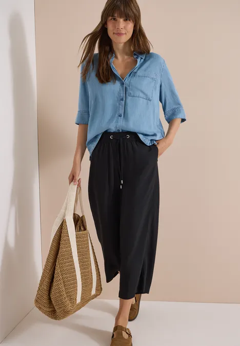 Frau trägt hellblaues Hemd, dunkle Culottes und Basttasche.