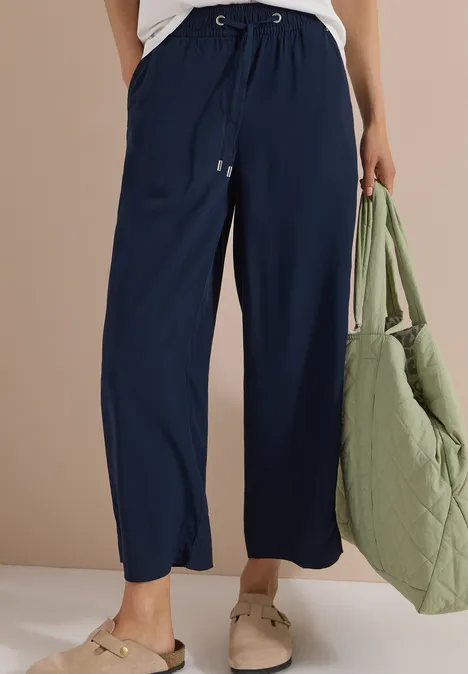 Weite, marineblaue Hose mit Kordelzug, kombiniert mit weißen Schuhen und grüner gesteppter Tasche.