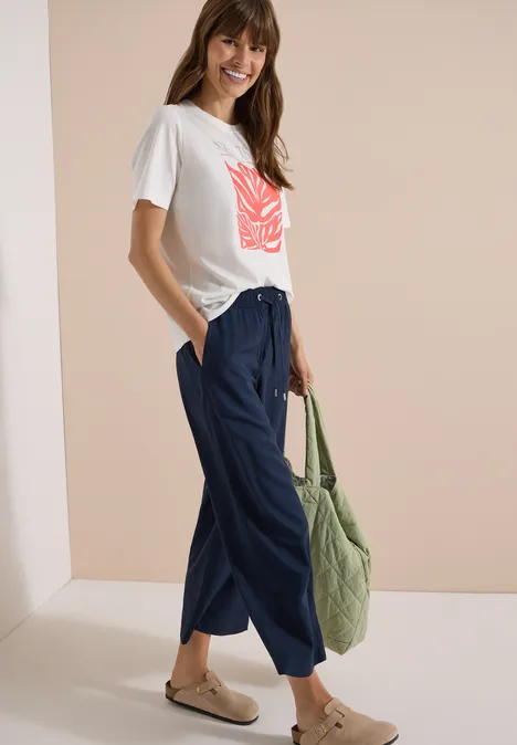 Frau in weißem T-Shirt mit rotem Print, blauen weiten Hose, Sandalen und grünem Shopper.