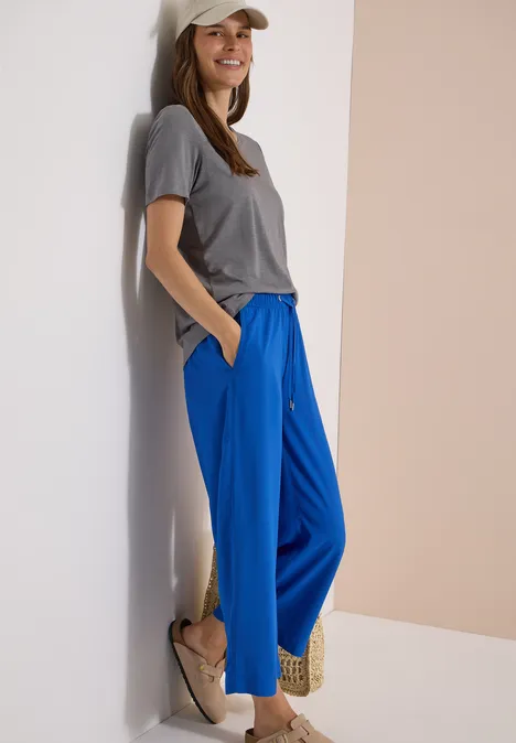 Frau lehnt an Wand, trägt graues T-Shirt, weite blaue Hose und beige Plateau-Schuhe.
