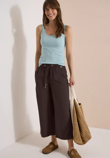 Frau trägt hellblaues Ärmelloses Top, schwarze Culotte, Basttasche und braune Sandalen vor beigem Hintergrund.