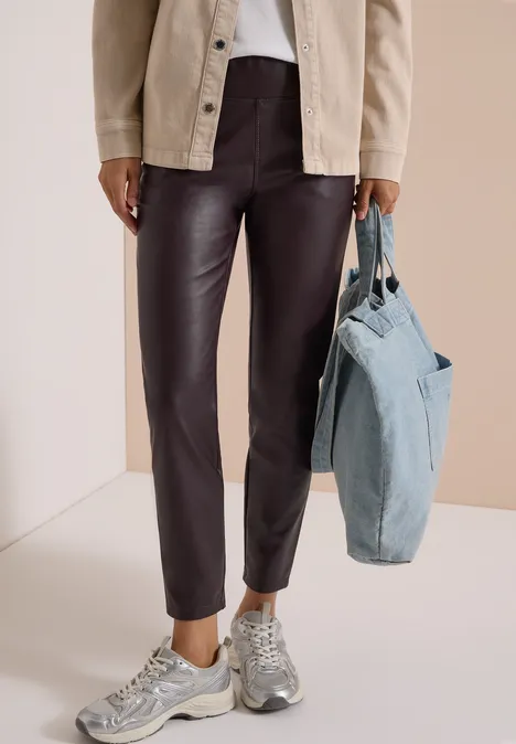 Een persoon draagt een bruine leren broek, grijze sneakers, een blauw denim tas en een lichte jas tegen neutrale achtergrond.
