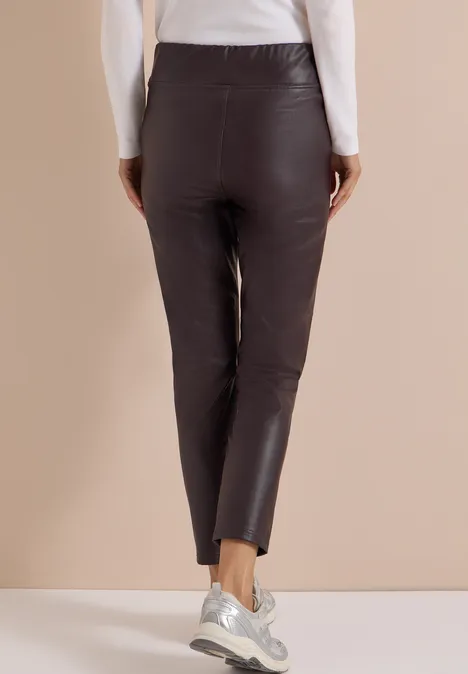 Achteraanzicht van persoon in zwarte, nauwsluitende leren broek en wit bovenstuk voor een beige achtergrond.