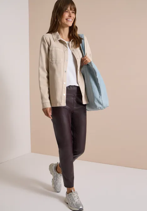 Vrouw met beige jasje, wit shirt, donkere legging, sneakers en grote lichtblauwe tas, glimlachend poseer.